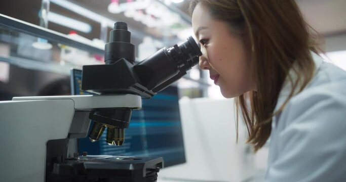 Medical Science Laboratory: Japanese Female Microbiologist Looking Through the Microscope, Analyzing Sample Genes and Writing Down Results on a Computer. Skillful Scientist Working in a Lab