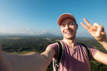 Happy man taking selfie photo from summer vacation day. Handsome tourist wearing cap and smiling at camera against tropical landscape. - 780517163