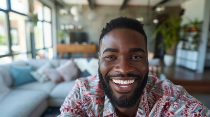 Smiling man with beard and mustache wearing patterned shirt in modern living room with large windows and greenery.