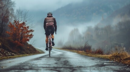 A solitary cyclist pedaling uphill on a misty mountain road surrounded by autumn foliage and a serene landscape. - obrazy, fototapety, plakaty