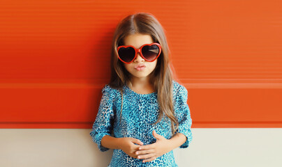 Beautiful little girl child in red heart shaped sunglasses and blue dress on city street