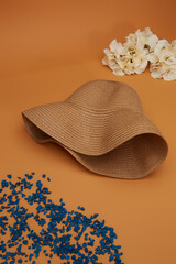 Close-up shot of a beige straw broad-brim sun hat. The casual straw hat with a wide brim is isolated on a brown background. Side view. Near the hat are white flowers and blue petals.