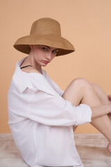 Close-up shot of a young woman in a beige straw hat with wide brim. The girl wearing a white shirt and a summer hat is sitting on the floor. The Model is posing on the beige background. Side view.