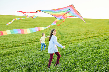 Smiling girl and brother boy running with flying colorful kites on the high grass meadow. Happy childhood moments