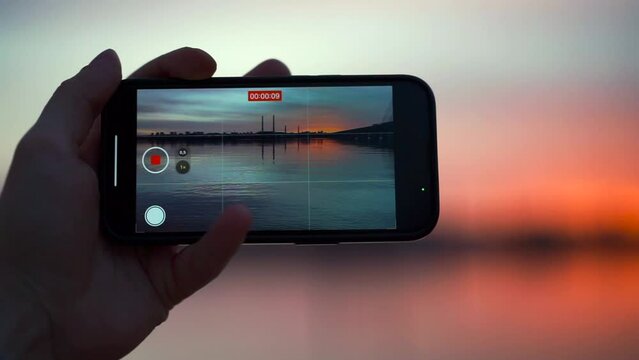 A man holds a phone in his hands and films an incredibly beautiful sunset over the city on the bank of a river that is filled with red from the sun, and the waves slowly roll onto the shore of the bea