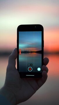 A man holds a phone in his hands and films an incredibly beautiful sunset over the city on the bank of a river that is filled with red from the sun, and the waves slowly roll onto the shore of the bea