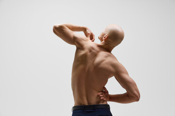 Rear view of sculpted physique of bald athletic man with beard in power stance against grey studio...