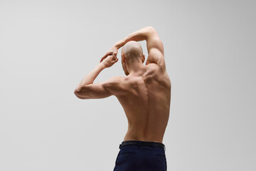 Rear view of sculpted physique of young bald man with beard in power stance against grey studio...