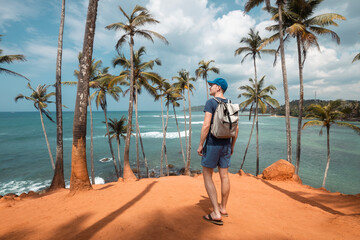 Traveler with backpack walking among coconut palm trees on hill aagainst tropical beach in Sri Lanka. .