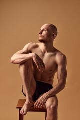 Portrait of young man with athletic body posing in underwear on stool looking away against sandy studio background. Concept of men's health, self care, fashion and beauty, healthy lifestyle. Ad - obrazy, fototapety, plakaty