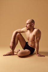 Young, toned bald man seated in introspective pose wearing black underwear against sandy studio...