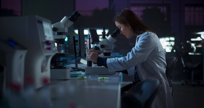 Medical Research and Development Laboratory: Hard-Working Asian Female Scientist Using Computer and Looking at a Sample in a Petri Dish Under a Microscope. Advanced Genetic Engineering Lab at Night