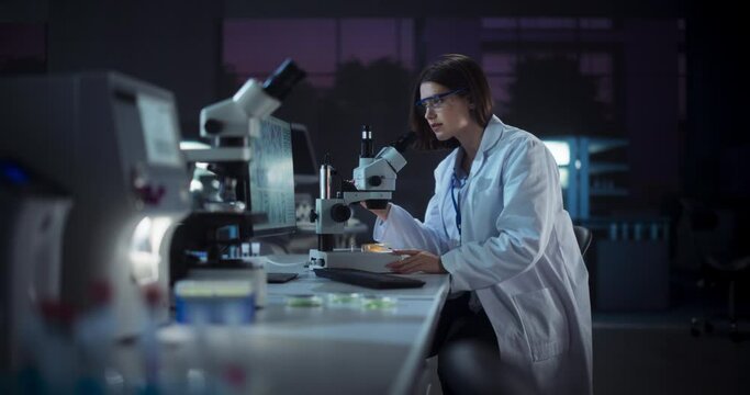 Medical Research and Development Laboratory: Hard-Working Female Scientist Using Computer and Looking at a Sample in a Petri Dish Under a Microscope. Advanced Genetic Engineering Lab at Night