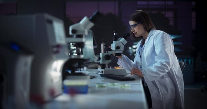 Cinematic Footage with a Beautiful Young Scientist Making an Important Innovative High Tech Discovery while Sitting in a Laboratory and Working with Microscope. Evening in Medical Research Center