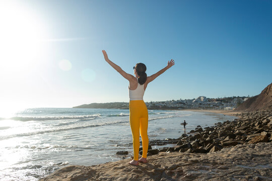 Sporty woman standing by the ocean with her arms raised enjoying the sun, positive emotions