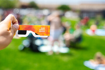 Side view of paper optics solar eclipse glasses scratch resistant polymer lenses filter harmful ultraviolet, infrared ray, blurry crow people on grassy yard watching totality show, Dallas, Texas