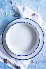 Handmade ceramic white plates with a blue stripe on the edge and empty seashells on blue concrete background. Top view.