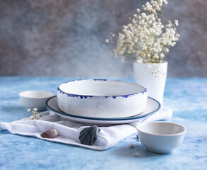Handmade ceramic white plates with a blue stripe on the edge, dried flowers and empty seashells on blue concrete background.