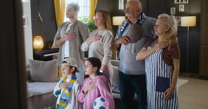 A multi-generational family of football fans stand in front of the TV and sing the national anthem before the start of the match