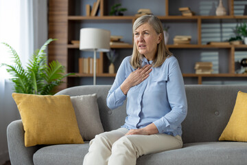 Senior woman touching chest while having pain in throat after coughing in domestic interior....