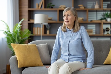 Stressed female in casual outfit sitting on grey sofa and looking aside with scared expression...