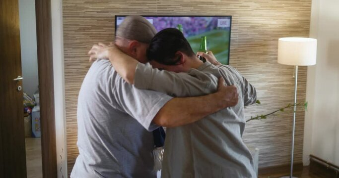 Multi-generational family of football fans jumps off the couch at the moment a goal is scored, celebrates and rejoices watching the match on TV at home. Senior man and his adult son celebrate a goal 