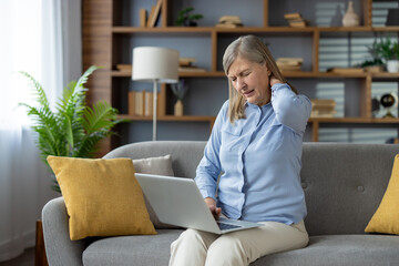 Tired mature lady in blue shirt rubbing tensed neck while sitting on couch with laptop on legs. Overloaded female touching back of head with closed wows for relieving strain in spinal muscles. - obrazy, fototapety, plakaty