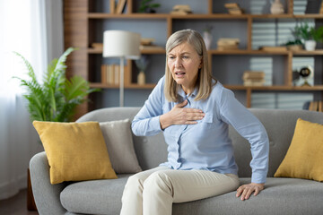 Elderly female grabbing chest with palm while sitting on pillowed couch with shocked expression....