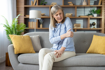 Mature woman having elbow injury and massaging sore spot with hand in stylish apartment interior. Unhealthy female in light clothes having tension in muscles while looking aside on hurting area. - obrazy, fototapety, plakaty
