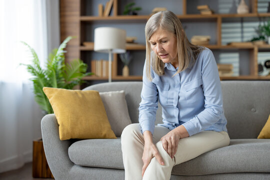 Pensioner woman sitting on grey couch and holding knee while bending over with grimace of pain. Suffering lady holding leg with inflammation related to ageing process and joint degeneration.