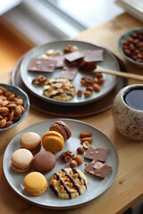 Plate with macarons, cookies, chocolate and nuts, cup of tea or coffee, book and reading glasses on the table. Selective focus. Colorful bookcase in the background.