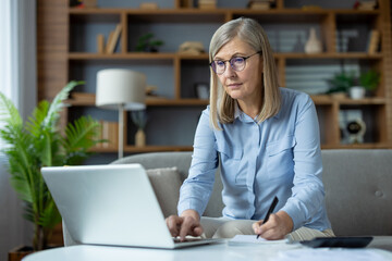 Stylish mature woman sitting on couch and using portable computer while writing in notebook at home. Attentive female joining remote classes for advanced training and making remarks from lecturer.