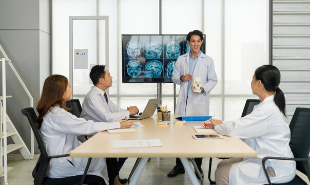 Young medical professional standing and presenting, holding a model of human skull and referencing image of brain scan displayed on a screen. The audience listening attentively with the presentation.
