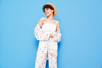 Young beautiful smiling female in trendy summer costume clothes. Carefree woman posing near blue wall in studio. Positive model having fun. Cheerful and happy. With curly short hairstyle, in hat