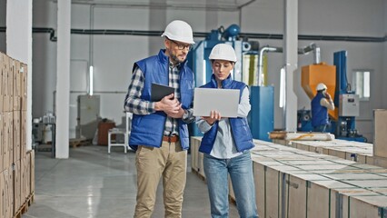 Two Caucasian workers wearing uniforms and helmets while standing in large storage or industrial...