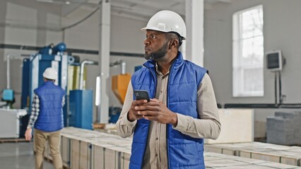 Zooming in on African American man wearing helmet while standing in middle of large facility. Specialist typing message on his phone. Considering production capacity while chatting with director.