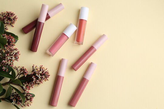 Different lip glosses and flowers on pale yellow background, flat lay. Space for text