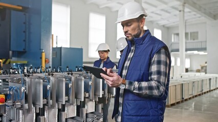 Side view of Caucasian man in safety helmet using tablet device while standing before manufacturing mechanism of foils. In background workers checking quality of productions. Production concept.