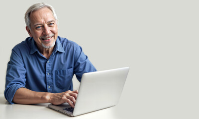 An older man in blue shirt sits at table with a laptop. Person on white background. Smile, gray hair, beard, mustache. Character in office workplace typing on laptop keyboard. Banner, flier, wallpaper - Powered by Adobe