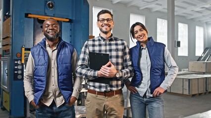 View of three multi-ethnic people smiling with joy while looking directly at camera. Handsome manager with clipboard standing between two engineers of manufacturing factory. Enjoying their work.