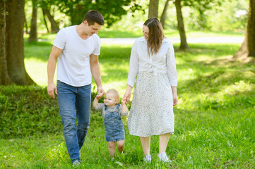 Fototapeta premium Toddler takes his first steps holding hands mom and dad in city park in the summer. Child learns walk independently using hands of his parents. Help with walking, motor skills and outdoor activities