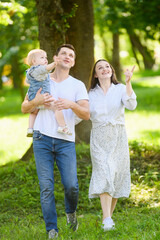 Fototapeta premium Parents and child go for walk in park, looking at everything around and talking, dad carries toddler in his arms. Family vacation concept, happy family moments, parenthood, walking in nature.Vertical