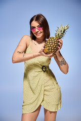 A brunette woman in a yellow dress joyfully holds a pineapple in a studio setting.