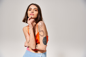A young woman with brunette hair and tattoos strikes a pose in a studio setting for a picture.