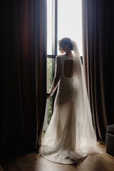 Elegant bride in a white dress standing near the window on her wedding day