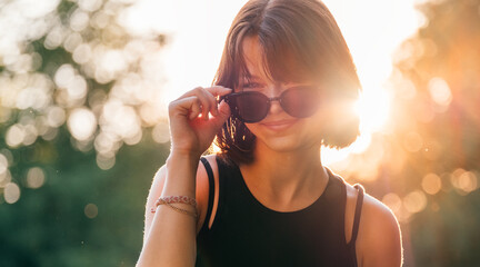 Beautiful young teenage girl in fancy sunglasses dressed in black dress challenging smiling at the camera with a warm sunset backlight. Beautiful people and a fashion concept image.