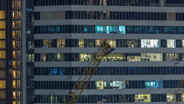 Office and residential buildings windows illuminated at night timelapse. Glass architecture, corporate building at evening with glowing lights. Cranes on a construction site in front