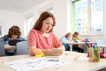 Red head little girl in glasses writing while sitting at the desk in classroom