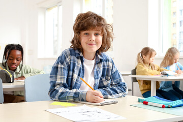 Smiling Caucasian schoolboy writing during the lesson, sitting at the desk while studying