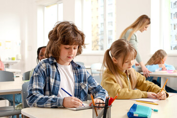 Schoolboy writing in notebook while sitting at the desk next to girl, teacher on the background